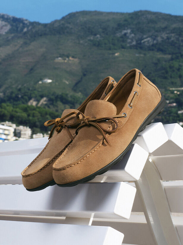 Men’s loafers with light-brown laces balanced on the back of a white bench with green hills in the background | GEOX | GEOX