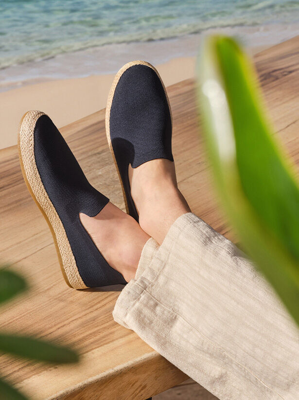 Man wearing navy-blue espadrilles and a lightweight stone-coloured pair of trousers sitting with his feet crossed and resting on a wooden table with the shoreline and sea in the background |GEOX | GEOX