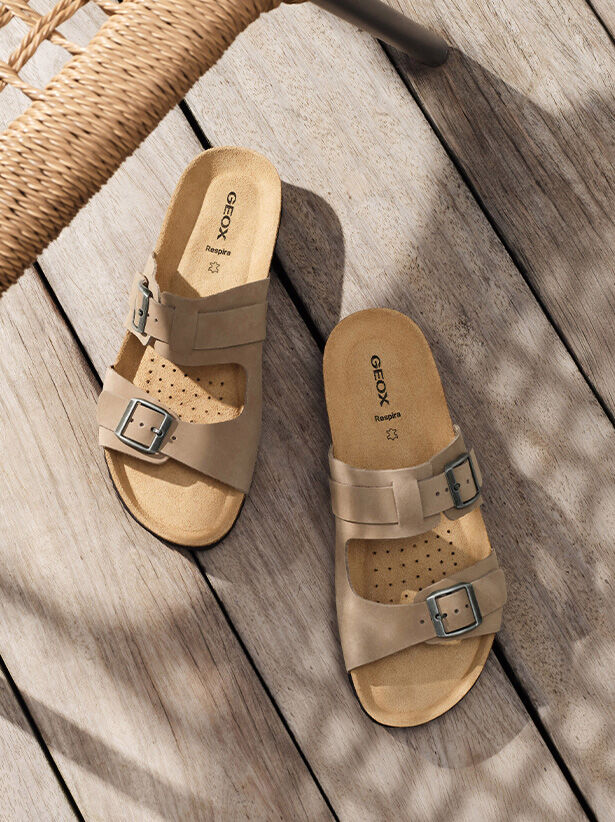 Taupe two-band sandals for men placed underneath a woven-straw chair on light-hued wooden flooring in the background |GEOX | GEOX