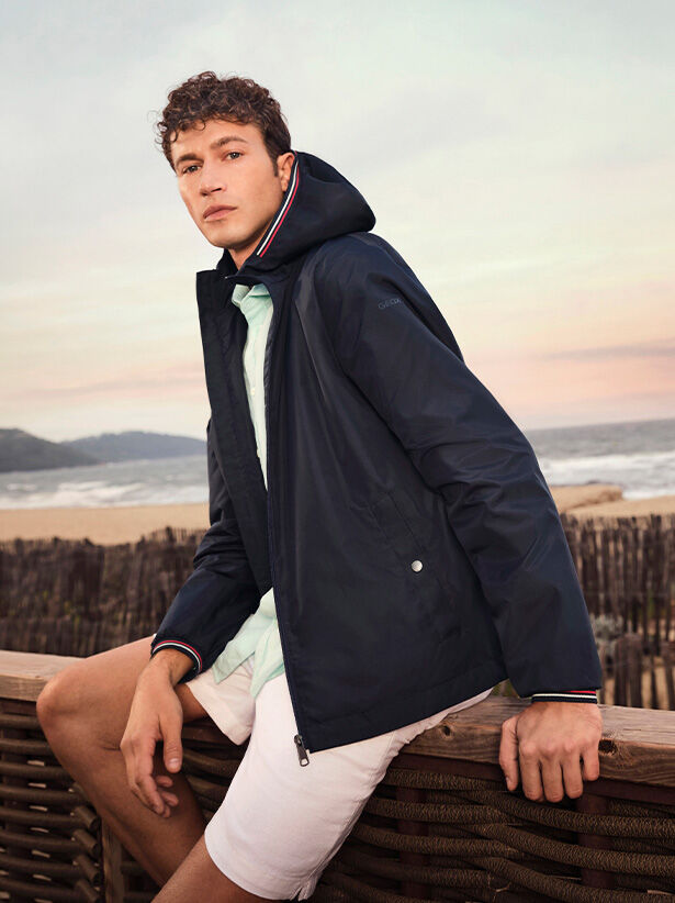 Man wearing a short hooded navy-blue jacket, white bermuda shorts and a mint-green shirt sitting on a wooden parapet with a beach scene in the background |GEOX | GEOX